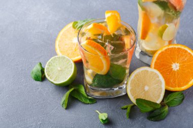 Limonlu içecekler buzla dolu bardaklarda servis edilir. Portakal, limon ve limon dilimleri gri bir masanın üzerindeki bardakların yanına konur. Taze nane yaprakları ekrana ekle.