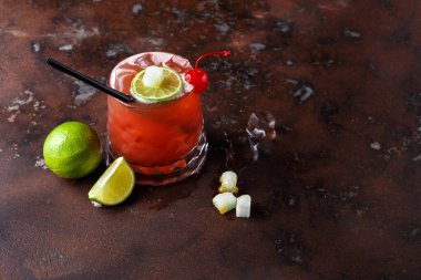 Parlak kırmızı bir kokteyl karanlık bir yüzeye oturur. Limon dilimi ve kirazla süslenmiş. Buz küpleri yakınlarda ve taze kireç parçaları içeceğin yanına yerleştirildi..