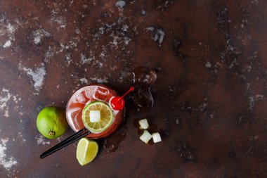 Bir kokteyl, içinde buz küpleri ve süslemeler olan karanlık bir yüzeye oturur. Limon ve bütün kireç yakınlarda. İçkinin üzerinde iki siyah pipetle kırmızı kiraz var..