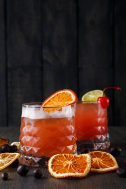 İki bardak buzlu, portakal dilimli ve kirazlı renkli içecek. Kurutulmuş portakal dilimleri ve koyu böğürtlenler, bardakları ahşap bir masanın etrafına sarmış. Bu bir akşam yemeği..