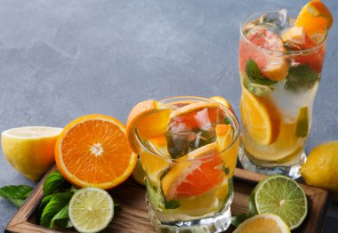 Turunçgil içecekleriyle dolu iki bardak ahşap bir tepsiye konur. İçeceklerde portakal, limon ve limon dilimleri var. Nane yaprakları ve buz küpleri görünür. Taze meyve bardakları sarıyor..