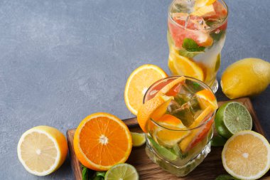 Renkli içeceklerle dolu iki bardak çeşitli narenciye meyveleriyle çevrili bir masada oturur. İçecekler arasında limon, limon ve taze görünüme eklenen portakal dilimleri de var..