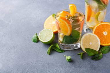 Gri bir yüzeye buz, nane yaprağı, portakal, limon ve limon dilimleri doldurulmuş iki bardak içki konur. Arkaplanda meyve karşıtlığının parlak renkleri.