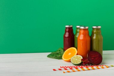 Meyve suyu şişeleri bir masanın üzerinde düzenlenir. Her şişe farklı bir renk. Taze meyve ve sebzeler şişelerin yanına oturur. Portakal dilimleri, limon dilimleri ve pancar kökü var..