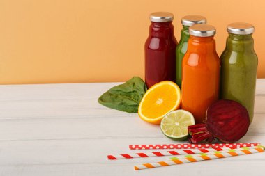 Dört şişe renkli meyve suyu ahşap bir masada duruyor. Yakında portakal, limon, ıspanak ve pancar gibi meyve ve sebzeler var. Çizgili pipetler sahneye biraz eğlence katar.