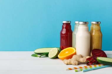 Meyve ve sebzelerle dolu bir masada farklı meyve suları bulunan şişeler bulunur. Kurulumda zencefil, salatalık, portakal ve limon gibi şeyler yer alıyor. Arka plan mavi..