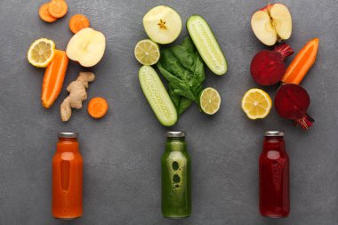 Taze meyve ve sebzeler üç şişe meyve suyu etrafında düzenlenir. Havuç, elma, salatalık ve pancar var. İçindekiler gri bir masada sağlıklı meyve suyu tarifleri öneriyor..