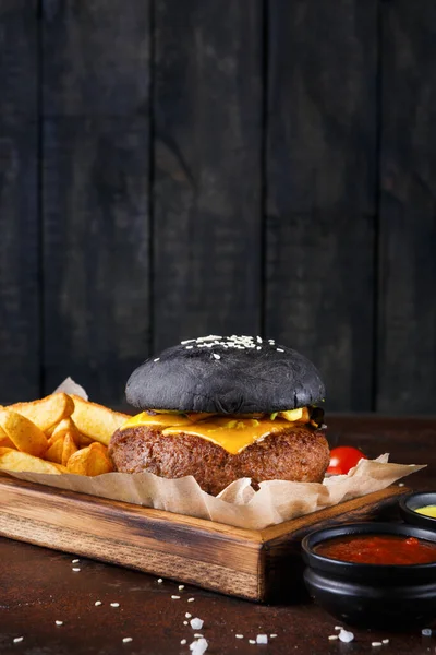 Peynirli bir siyah burger, altın kızartmalarla çevrili tahta bir tabakta kâğıt üzerinde oturur. Tabağın yanına küçük sos kaseleri konur. Arkaplanda koyu tahta paneller var..