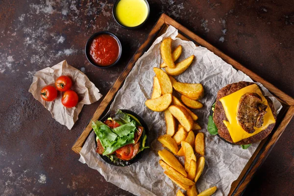 Peynirle kaplı bir hamburger, ahşap tepside kızarmış patateslerin yanında oturur. Yeşillik ve domatesli bir salata da dahil. Soslar yakınlardaki küçük kaselerde sergileniyor..