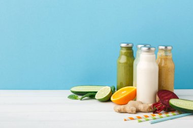 Dört farklı meyve suyu şişesi bir masaya konur. Salatalık, zencefil ve limon gibi taze meyve ve sebzeler mavi arka plandaki şişelerin yanına dizilir..