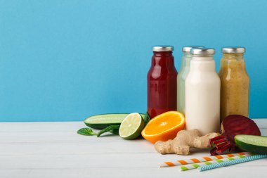 Farklı meyve sularından şişeler bir masada oturur. Limon, portakal, salatalık ve zencefil gibi taze meyve ve sebzeler şişelerin etrafını sarar. Kamışları içkilerin yanına koyun..