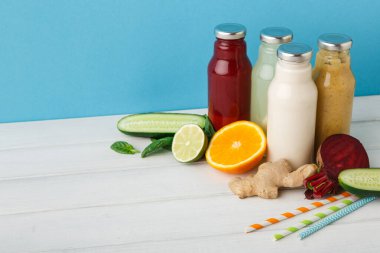 İçeceklerle dolu birkaç cam şişe beyaz bir ahşap masaya yerleştirilir. Taze meyve ve sebzeler şişelerin yanında düzenlenir. Limon, salatalık ve portakal dilimleri görünür..