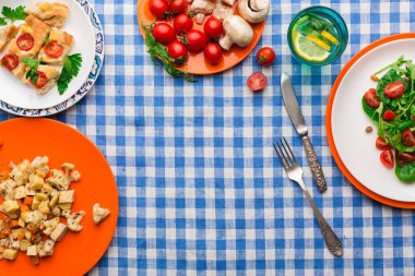 Farklı tabaklarda salata, pişmiş sebze ve taze domates gibi çeşitli yiyecekler bulunur. Limonlu bir içecek mavi ve beyaz bir masa örtüsünün üzerinde duruyor..
