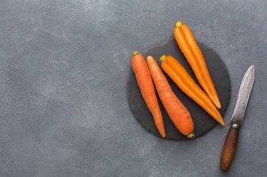 Havuçlar, yanlarında bir bıçakla yuvarlak bir taş tahtaya konur. Havuçlar parlak turuncudur ve doğal bir şekli vardır. Ayarlar, mutfak yüzeylerinin tipik gri bir arka planına sahiptir..