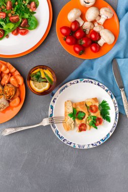 İçinde domatesli salata, bir tabak sebze ve beyaz tabakta ana yemek olan bir masa seti. Limon dilimli bir içecek de mevcut..