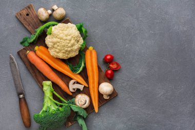 Taze sebzeler arasında karnabahar, havuç, brokoli ve tahta kesme tahtasına yerleştirilmiş mantarlar yer alıyor. Gri arka planı olan sebzelerin yanında bir bıçak duruyor..