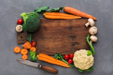 Düz bir yüzeye tahta kesme tahtası yerleştirilir. Çevresinde brokoli, karnabahar, havuç, vişneli domates ve mantar gibi çeşitli taze sebzeler vardır..