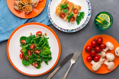 Masadaki renkli tabaklara farklı yemekler yerleştirilmiştir. Yeşillik ve domatesli bir salata var. Diğer tabaklar arasında pişmiş yemek ve limonlu içecek yer alıyor..