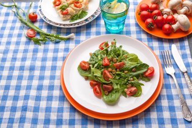 Taze yeşillik ve kiraz domatesinden yapılmış bir salata tabakta. Yakınlarda, kareli bir masa örtüsünün üzerinde atıştırmalıklar ve içecekler var. Ayar parlak ve davetkar görünüyor..