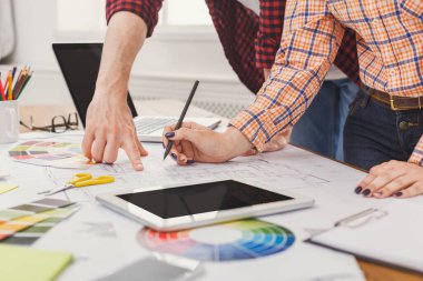 İki kişi tasarım planları ve renk örnekleriyle dolu bir masaya odaklanmış durumda. Biri bir planı işaret ederken, diğeri de notlar yazıyor. Cihazlar ve cihazlar etraflarında..