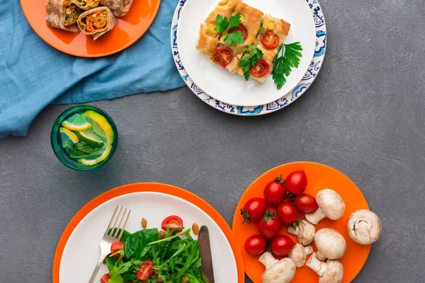 Üç tabakta sebzeli bir salata, küçük domatesler ve mantarlar da dahil olmak üzere çeşitli yiyecekler yer alıyor. Limon dilimli bir içecek yakınlarda durur. Ayar gri bir masada, üzerinde mavi bir bez var..