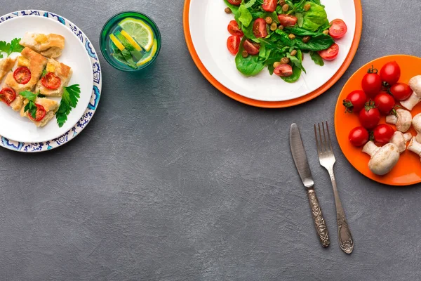 Masada domatesli ve yeşillikli bir salata, fırınlanmış bir tabak ve taze mantarlar da dahil olmak üzere çeşitli yiyecekler sergilenmektedir. Limon dilimli bir içecek de görülebilir..