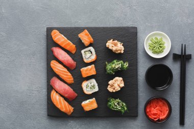 Ahşap tahta, nigiri ve rulo da dahil olmak üzere farklı suşi türleri barındırır. Ayrıca wasabi, soya sosu ve dilimlenmiş zencefilli kaseler de var. Çubuklar tahtanın yanında..
