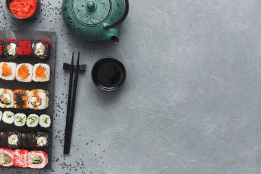 Bir tabak suşi rulosu gri bir masada bir çaydanlık ve küçük bir fincanın yanında duruyor. Soya sosu ve wasabi yakınlarda. Yemek çubukları kullanıma hazır hale getirilir..