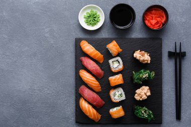 Siyah tabakta somon, ton balığı ve rulo da dahil olmak üzere çeşitli suşi türleri bulunur. Küçük kaseler dolusu wasabi, soya sosu ve turşulu zencefil tabağın yanına otur. Yemek çubukları yakınlarda dinleniyor..