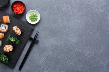 Bir suşi tabağı siyah bir taş tahtada duruyor. Farklı suşi türleri düzgün bir şekilde düzenlenir. Çubuklar tabağın yanında duruyor. Yanında soya sosu, wasabi ve zencefil turşusu var..