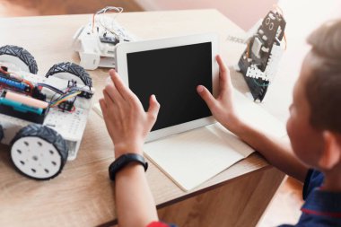 Bir çocuk elinde tablet olan bir masada oturuyor. Bir robot araba ve diğer oyuncak robotlar yakınlarda. Çocuk robotları kontrol etmek veya programlamak için tableti kullanmaya odaklanmış görünüyor.