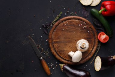 Tahta kesme tahtası, etrafında taze sebzeler olan karanlık bir yüzeyde oturur. Yemek pişirmek için bıçak, mantar, domates, kabak ve dolmalık biber var. Sahne yemek hazırlığı için hazırlandı..