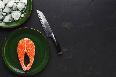 Bir somon filetosu yeşil bir tabakta bir kase buzun yanında oturur. Yakınlarda keskin bir bıçak var. Karanlık yüzey bir mutfak ortamını işaret ediyor. Deniz ürünleri hazırlıklarına odaklanın..