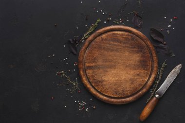 Tahta kesme tahtası, çeşitli otlar, baharatlar ve bir bıçakla çevrili karanlık bir yüzeyde oturur. Yemek ya da yemek hazırlamak için hazırlanmış organize bir sahne..