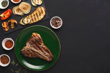 Bir biftek yeşil bir tabakta ızgara sebze ve baharat kaselerinin yanında oturur. Dekor karanlık, yemeğin detaylarını vurguluyor..