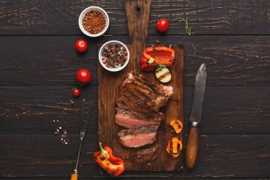 Izgara biftek dilimleri ahşap kesme tahtasının üzerinde duruyor. Yakınlarda taze sebzeler ve baharatlar var. Yemek hazırlama ya da akşam yemeği deneyimi öneriliyor..