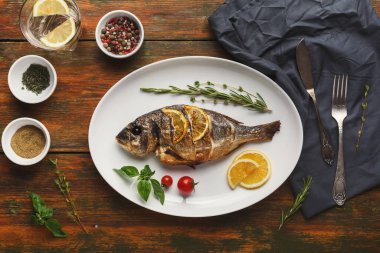 Bütün bir balık, limon dilimleri ve otlarla birlikte beyaz bir tabakta düzenlenir. Tabağın etrafında baharatlı kaseler ve ahşap bir masa üzerinde bir bez var..