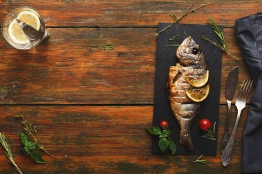 Pişmiş bir balık, limon dilimleriyle siyah bir tabakta sergilenir. Sofrada bir bardak içki, şifalı otlar ve kırsal ahşap bir masa üzerindeki kiraz domatesleri yer alıyor..