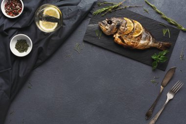 Izgara bir balık, üzerinde limon dilimleri olan siyah bir tahta üzerine konur. Orada otlar, baharatlar ve bir bardak su var..