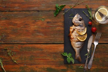Izgara bir balık, limon dilimleriyle siyah bir tabakta oturur. Taze bitkiler ve vişneli domatesler tabağın etrafını sardı. Çatal bıçak takımı tabağın yanına konur. Bu sahne ahşap bir masada geçiyor..