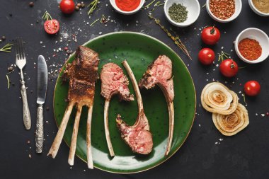 Kuzu pirzolası yeşil bir tabağa konur. Tabağı çevreleyen çeşitli baharatlar ve taze malzemeler. Sahne karanlık bir yüzeye kurulmuştur. Kırmızı domates ve bitkiler var..