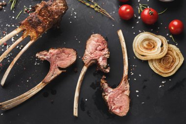 Bir koleksiyon, koyu bir yüzeyde sıralanmış ızgara et parçalarını gösteriyor. Taze sebzeler ve otlar etin etrafını sarar. Ortam yemek hazırlama ve evde yemek pişirmeye odaklanıyor..