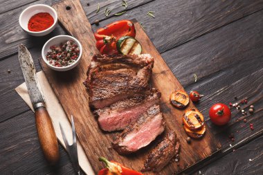 Izgara sebze ve baharatla birlikte ahşap bir tahtanın üzerinde ızgara biftek dilimleri koleksiyonu. Sahne kırmızı biber ve domateslerle servis edilen bir yemek düzenini gösteriyor..