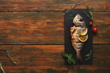 Pişmiş bir balık, karanlık bir servis tahtasına yerleştirilir. Taze bitkiler ve vişneli domatesler balığı çevreledi. Ahşap masa sıcak bir dokunuş ekliyor. Bu ortam sıradan bir yemek deneyimi olduğunu gösteriyor..