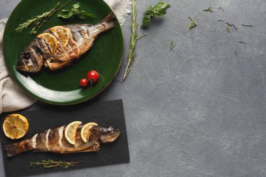 İki ızgara balık, yeşil bir tabak ve siyah bir tahta üzerinde sergileniyor. Taze otlar ve domatesler mutfaktaki balıkları çevreledi. Sahne, servise hazır bir yemeği ön plana çıkarıyor..