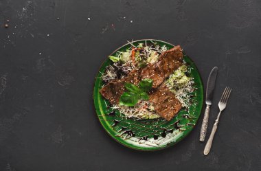 Üzerinde et dilimleri, taze salata ve fesleğen bulunan yeşil bir tabakta servis edilir. Ayar, karanlık bir yüzeyde tabağın yanına yerleştirilmiş çatal bıçakları içeriyor..