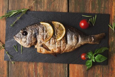 Koca bir balık siyah bir tabakta pişirilip servis edildi. Balık limon dilimleri, kiraz domatesleri ve taze otlarla süslenir. Ayar, tahta masa yüzeylerini gösteriyor..