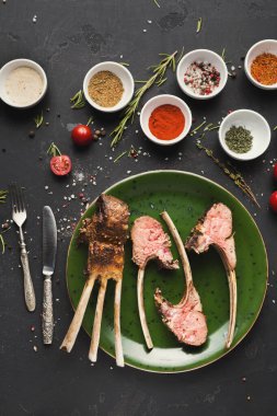 Kuzu pirzolası yeşil bir tabakta duruyor. Etrafı çeşitli baharatlarla çevrili. Taze bitkiler ve kiraz domatesleri masaya saçıldı. Hazırlıklar için araç gereçler yakına yerleştirilir.