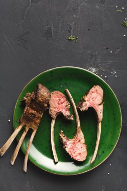 Izgara kuzu pirzolası yeşil bir tabağa konur. Et dışarıdan pişiyor ve içinde pembe bir renk var. Tabak karanlık bir yüzeye yerleştirilmiş..