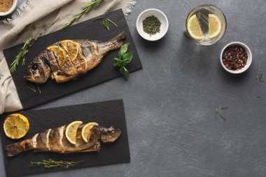 İki ızgara balık, limon dilimli siyah tabağa konur. Taze otlar ve baharatlar yakınlardaki kaselerde sergilenir. Mutfak ortamında yemek hazırlığı yapılıyor..